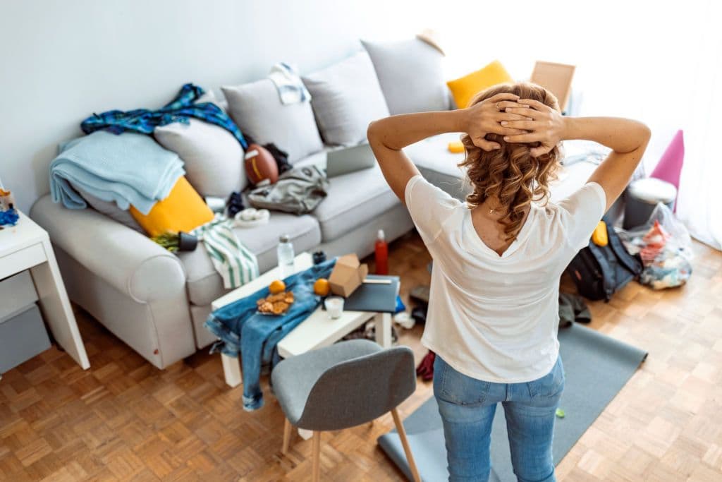 Vrouw staat met handen in haar haar in een rommelige woonkamer vol spullen